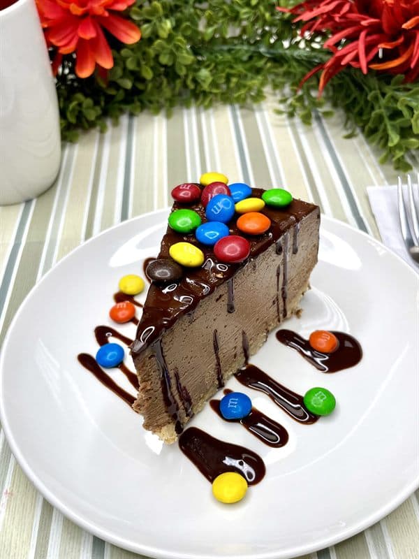 A slice of chocolate cheesecake piled with candy-shell chocolates and finished with chocolate drizzle.